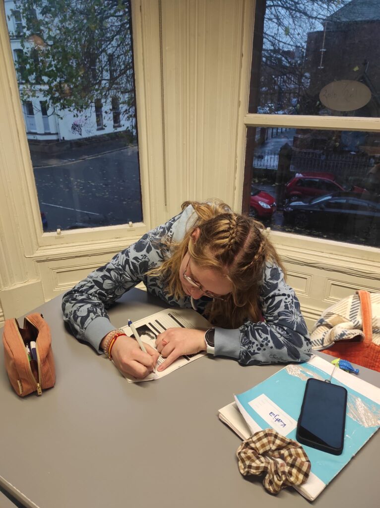 A female student, with blonde hair and glasses, wearing a floral hoodie , sitting at a desk at IH Belfast, holding a pen, filling in a handout. 