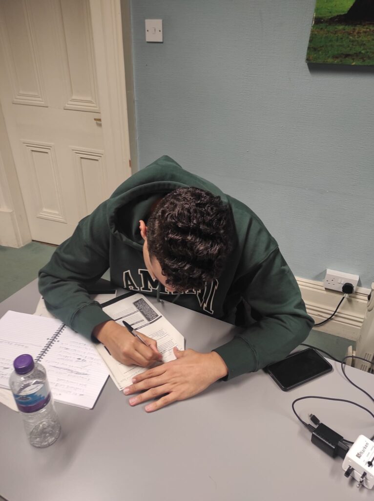 A male student, with black hair, wearing a green hoodie. He's sitting at the desk, holding a black pen, and filling in a handout. 
